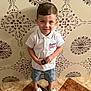 child, boy, standing, white_polo, denim_shorts, nike_sandals, tile_floor, patterned_wall, smiling, short_hair, indoor, casual_clothing, holding_object, footwear, portrait, happy, cute, playful, young, person