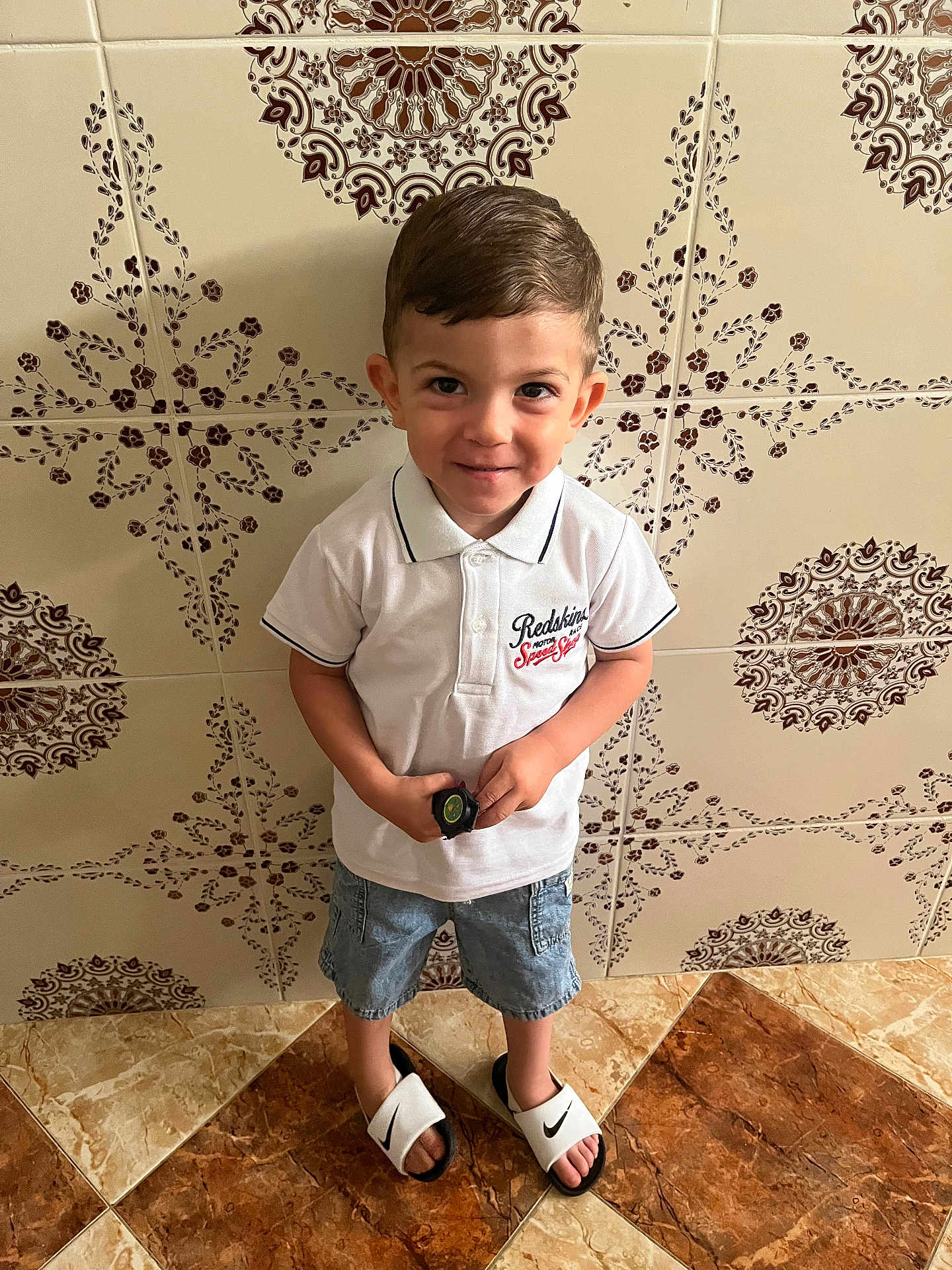 Iyad participe au concours pour gagner de l'argent avec cette photo : child, boy, standing, white_polo, denim_shorts, nike_sandals, tile_floor, patterned_wall, smiling, short_hair, indoor, casual_clothing, holding_object, footwear, portrait, happy, cute, playful, young, person
