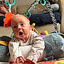 baby, infant, child, play_mat, toy, stuffed_animal, person, couch, home_interior, expression, wide_eyes, open_mouth, hand, sleeve, patterned_clothing, background, seated, colorful, indoor, curious