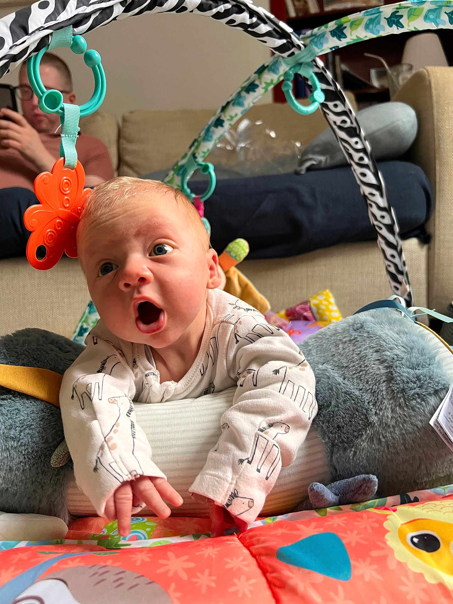 Aidan joined the competition — help win amazing prizes! baby, infant, child, play_mat, toy, stuffed_animal, person, couch, home_interior, expression, wide_eyes, open_mouth, hand, sleeve, patterned_clothing, background, seated, colorful, indoor, curious