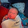 Emerson is registered to the contest to win money with this photo: baby, infant, blanket, bed, pillow, red_clothing, wrapped, surprised_expression, face, person, indoors, sleeping_area, patterned_bedspread, floral_pillow, cute, newborn, child, lying_down, soft_lighting, cozy