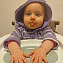 baby, child, hood, purple_clothing, high_chair, tongue_out, hands, face, blue_eyes, indoor, sitting, playful, sweater, infant, person, cute, portrait, table, expression, young_child