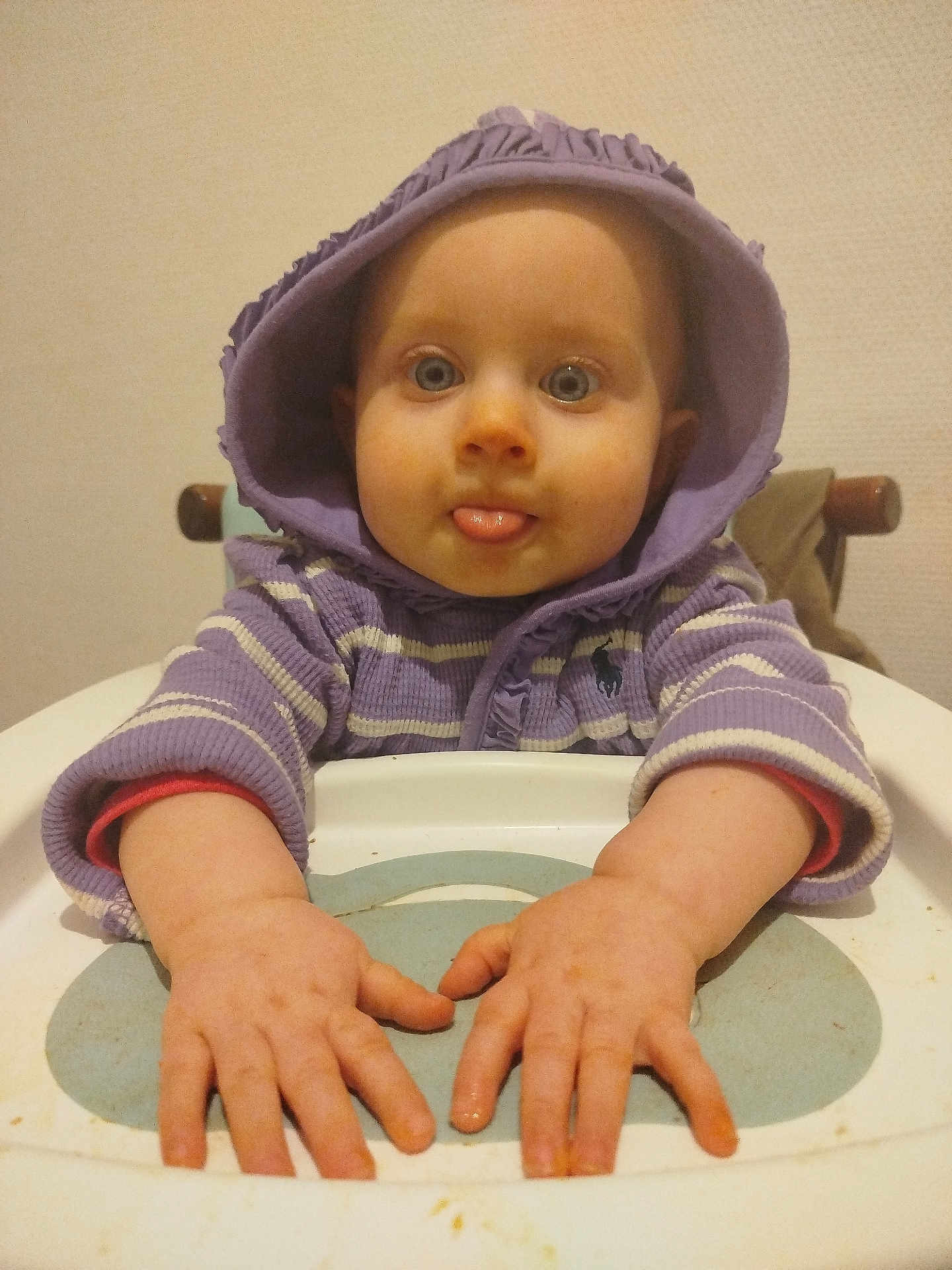 Louve participe au concours pour gagner de l'argent avec cette photo : baby, child, hood, purple_clothing, high_chair, tongue_out, hands, face, blue_eyes, indoor, sitting, playful, sweater, infant, person, cute, portrait, table, expression, young_child