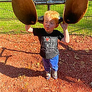 Clint Jr is registered to the contest to win money with this photo: child, playground, outdoor, grass, fence, red_hair, tshirt, shoes, shadow, woodchips, pond, sunny, curious, smudged_face, play_equipment, blue_pants, person, daylight, fun, nature