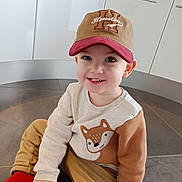 Noah participe au concours pour gagner de l'argent avec cette photo : toddler, child, cap, sweater, fox_design, red_socks, pants, floor, kitchen, cabinet, smile, sitting, cute, indoors, portrait, happy, young_child, clothing, person, casual_wear