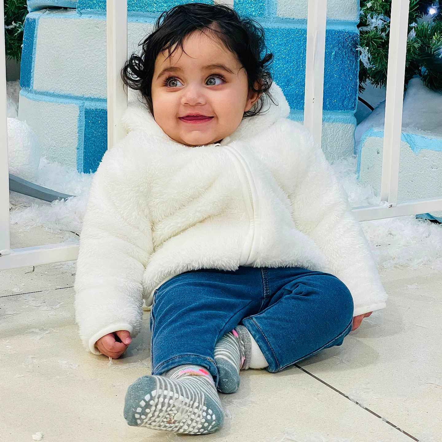 Ibadat is registered to the contest to win money with this photo: background, blue_jeans, child, curly_hair, cute, face, feet, floor, greenery, happy, holiday_theme, indoor, person, playful, sitting, smiling, socks, toddler, white_jacket, winter_decor