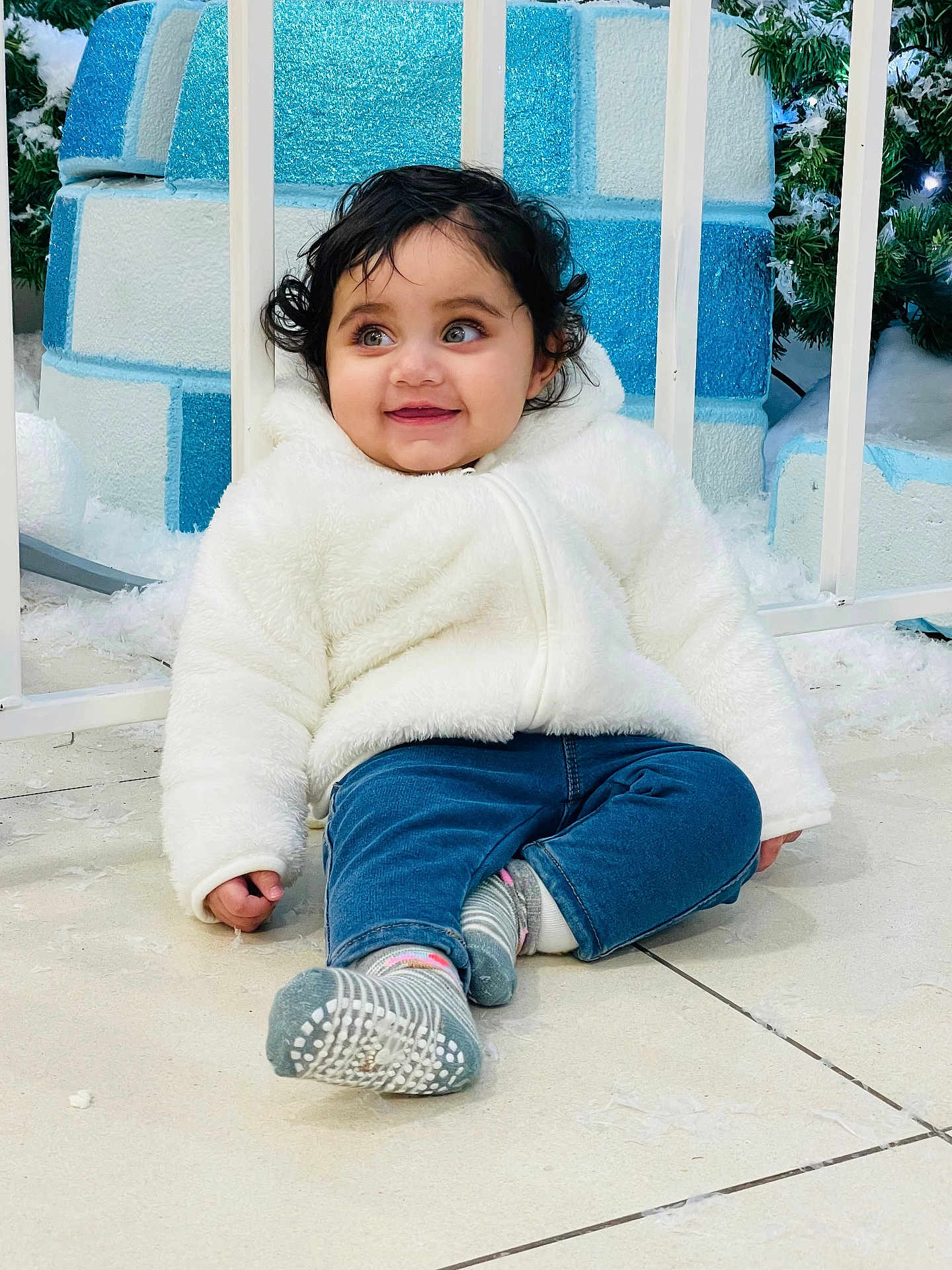 Ibadat is registered to the contest to win money with this photo: toddler, child, smiling, white_jacket, blue_jeans, sitting, floor, indoor, winter_decor, holiday_theme, curly_hair, happy, cute, face, person, feet, socks, background, greenery, playful