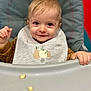 baby, child, high_chair, bib, food, snack, smile, face, hands, clothing, indoor, person, cute, young_child, feeding, expression, seat, table, playful, portrait