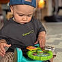 César participe au concours pour gagner de l'argent avec cette photo : toddler, child, toy, indoor, cap, blue_cap, striped_shorts, black_shirt, play, floor, concentration, cute, baby, hand, sitting, colorful_toy, young_child, person, casual_clothing, soft_background