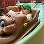toddler, child, green_cap, boat_ride, water_ride, amusement_park, brown_boat, outdoor, curious, water, playful, fun, young_child, casual_clothing, fence, wooden_furniture, summer, daytime, family_fun, portrait
