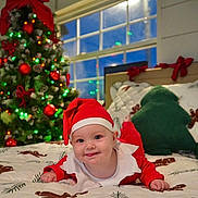 Melody is registered to the contest to win money with this photo: baby, bedspread, child, christmas, cute, decorations, festive, green_pillow, holiday, holiday_season, indoors, infant, lights, pillow, red_clothing, santa_hat, smile, smiling, tree, window