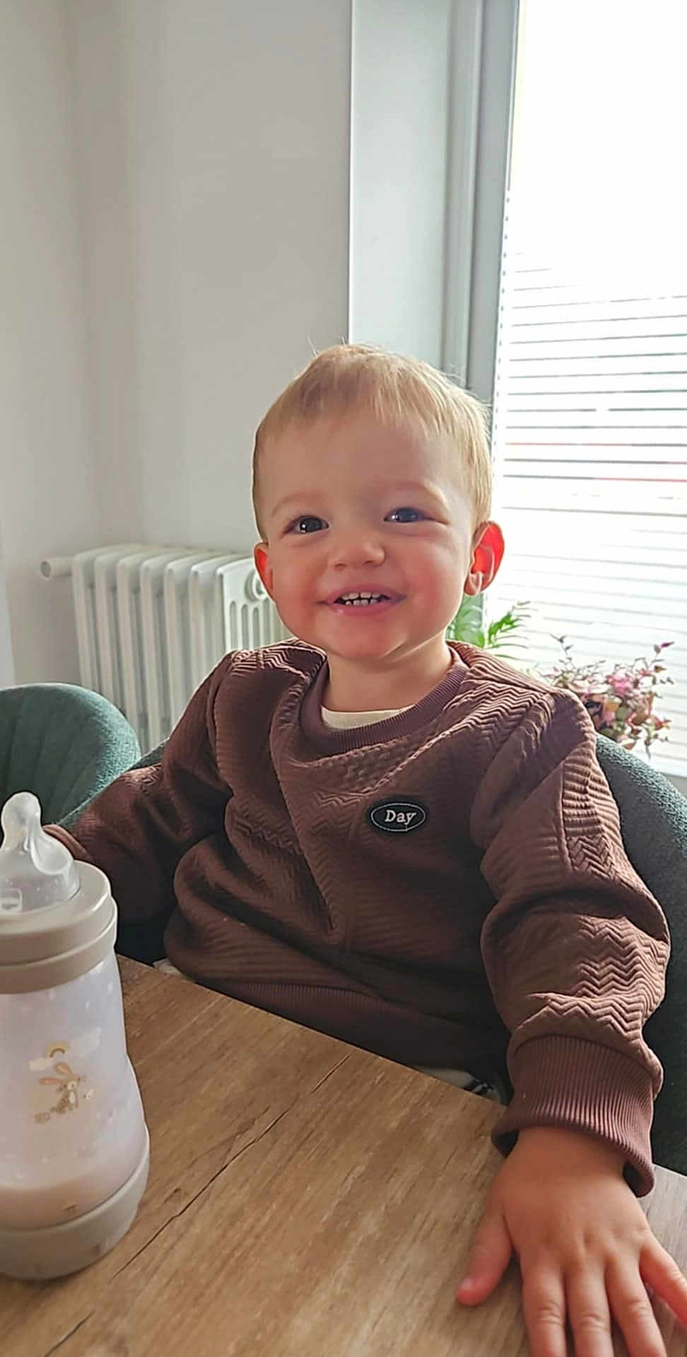Tommy a rejoint le concours — aidez-le/la à gagner de superbes lots ! toddler, child, smiling, baby_bottle, brown_sweater, table, indoor, plant, window, blinds, radiator, hand, face, person, happy, seated, cozy, casual_clothing, home, daylight