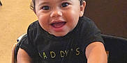 Kaiden is registered to the contest to win money with this photo: toddler, child, smiling, indoor, table, black_shirt, curly_hair, happy, face, hand, person, furniture, wood, seated, young_child, cute, portrait, bright_eyes, candid, casual