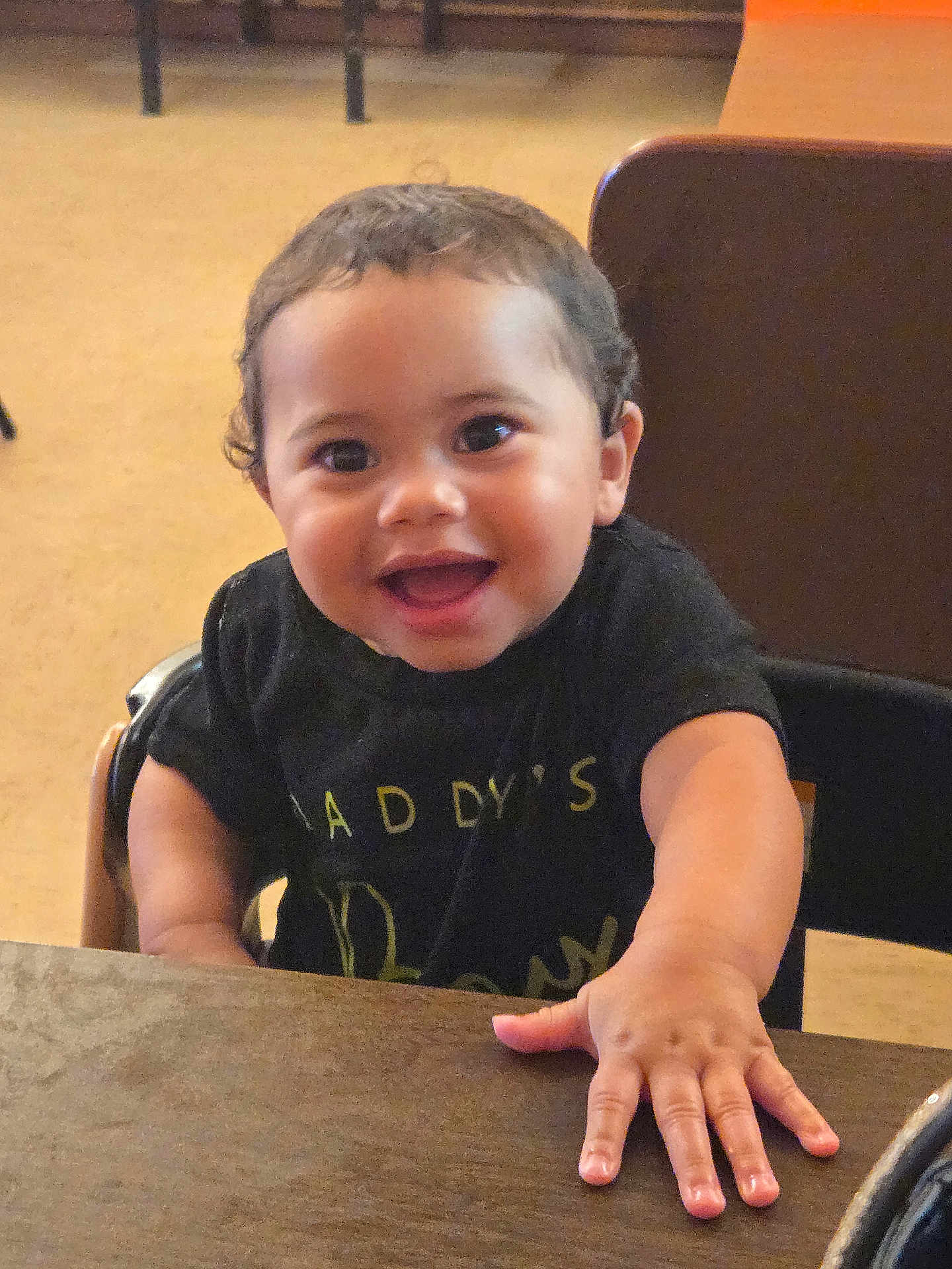 Kaiden is registered to the contest to win money with this photo: toddler, child, smiling, indoor, table, black_shirt, curly_hair, happy, face, hand, person, furniture, wood, seated, young_child, cute, portrait, bright_eyes, candid, casual