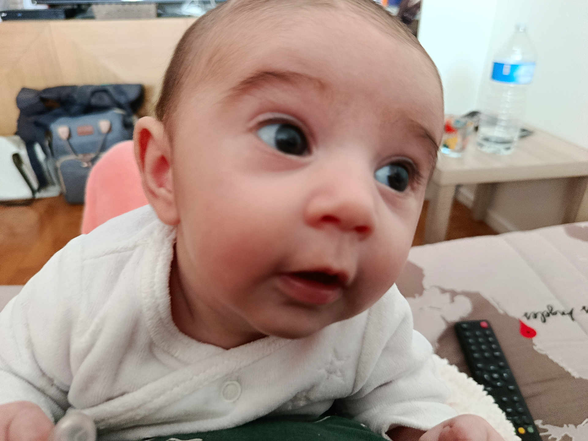 Ellie a rejoint le concours — aidez-le/la à gagner de superbes lots ! baby, infant, close_up, big_eyes, surprised_expression, face, eyes, mouth, ear, cheek, onesie, indoor, bedroom, water_bottle, remote_control, table, bag, blanket, portrait, cute