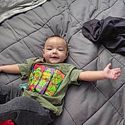 Ramon is registered to the contest to win money with this photo: baby, child, tshirt, green_tshirt, cartoon, ninja_turtles, bedspread, gray_bedspread, blanket, pillow, lying_down, smiling, arms_outstretched, indoors, cozy, clothing, person, cute, infant, happy