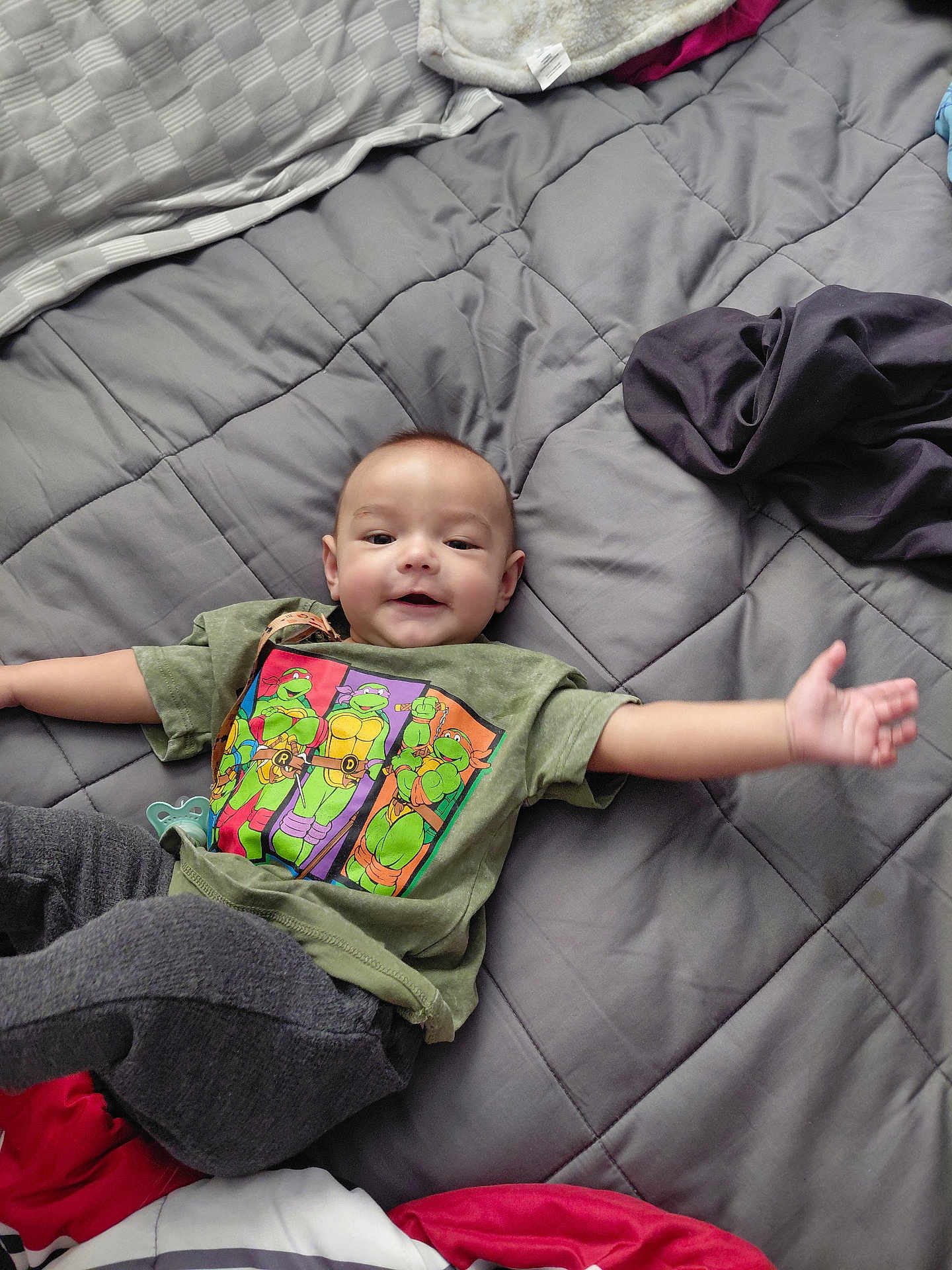 Ramon is registered to the contest to win money with this photo: baby, child, tshirt, green_tshirt, cartoon, ninja_turtles, bedspread, gray_bedspread, blanket, pillow, lying_down, smiling, arms_outstretched, indoors, cozy, clothing, person, cute, infant, happy