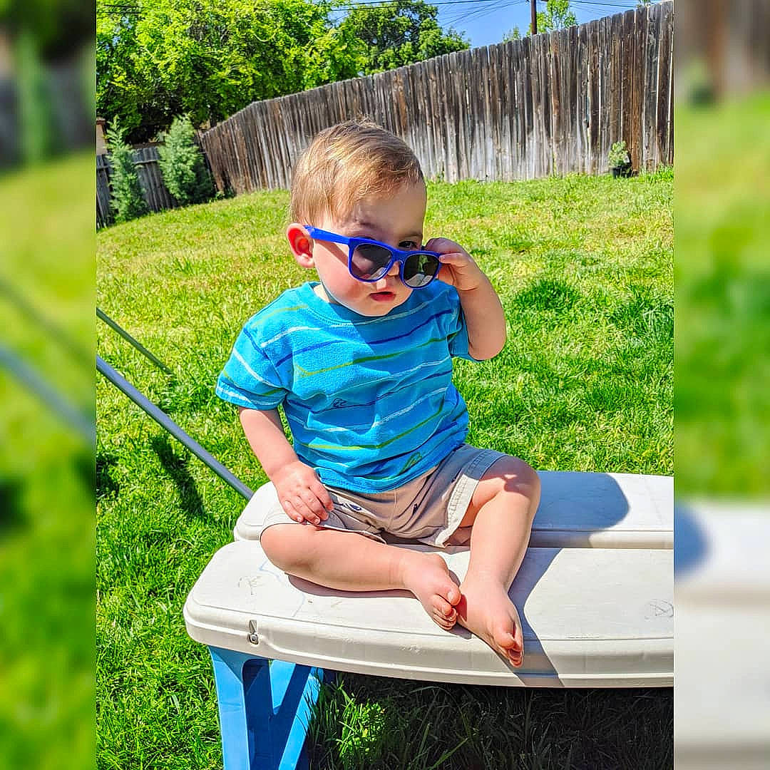 Dominik is registered to the contest to win money with this photo: child, cool, eyewear, furniture, glasses, grass, leisure, outdoor_furniture, person, plant, play, sitting, smile, spring, summer, sunglasses, t_shirt, table, toddler, turquoise