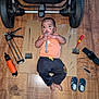 baby, child, floor, wooden_floor, tools, wrench, screwdriver, stroller_wheel, cardboard, black_marks, clothing, pants, orange_shirt, barefoot, footwear, toy_shoes, indoor, person, infant, playful