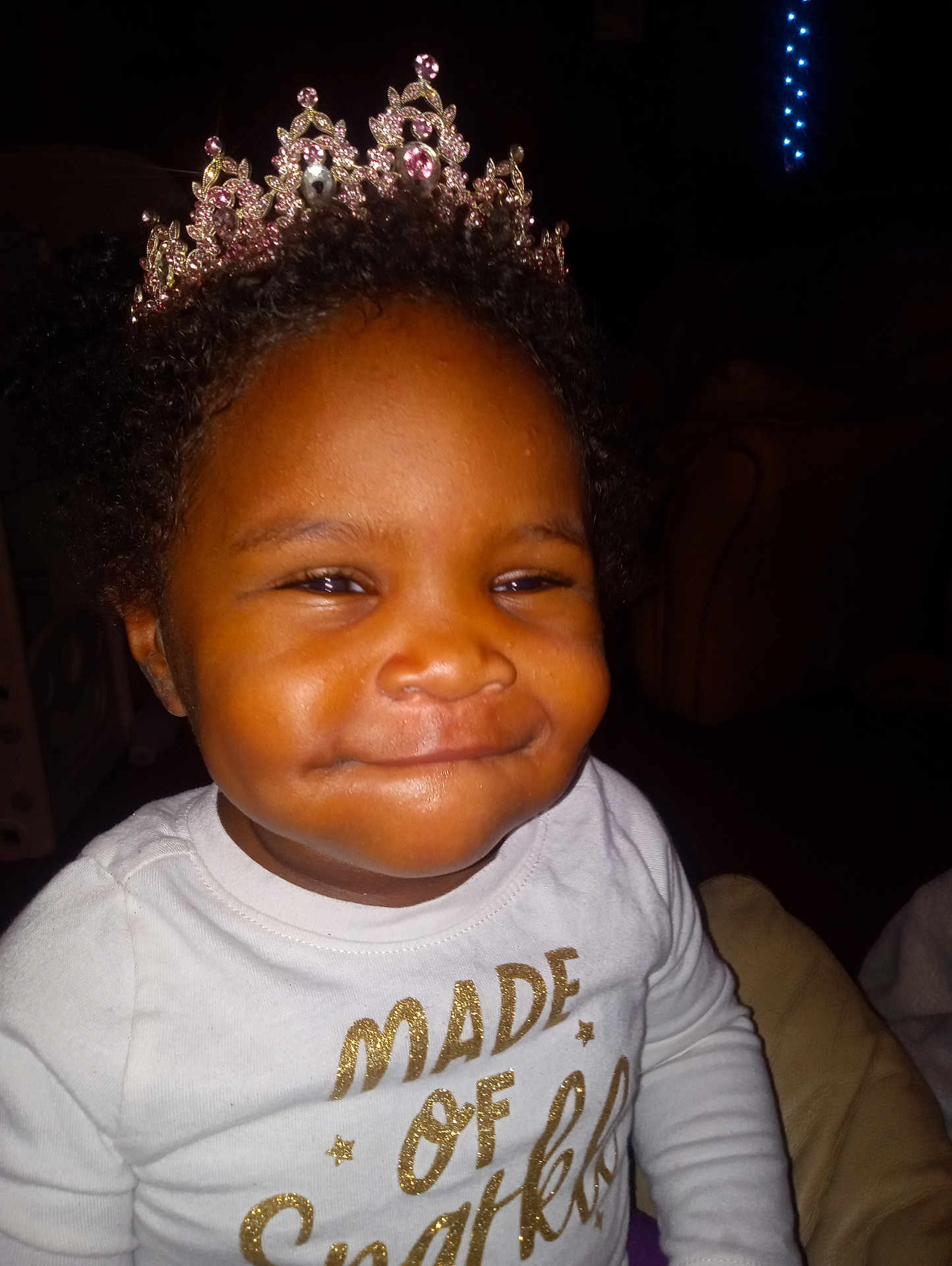 A'Kayda is registered to the contest to win money with this photo: child, toddler, tiara, crown, smile, portrait, closeup, indoor, dark_background, shirt, gold_text, glitter, curly_hair, happy, baby, living_room, cozy, cute, sparkle, flash_photography