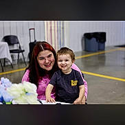 Langston joined the competition — help win amazing prizes! child, adult, smile, indoor, table, flowers, chair, trash_can, suitcase, concrete_floor, wall, portrait, family, hugging, happy, person, clothing, short_hair, event_space, folding_chair