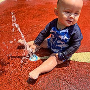 Lawton joined the competition — help win amazing prizes! toddler, child, water, splash, playground, wet_surface, swimwear, blue_clothing, red_ground, sunlight, outdoor, summer, play, baby, barefoot, sitting, joyful, water_fountain, skin, head