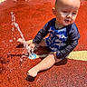 toddler, child, water, splash, playground, wet_surface, swimwear, blue_clothing, red_ground, sunlight, outdoor, summer, play, baby, barefoot, sitting, joyful, water_fountain, skin, head