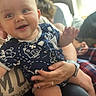 adult_hand, baby, blue_clothing, blurred_background, candid, casual, child, face, happy, holding, indoor, infant, onesie, person, portrait, ring, seated, smiling, watch, window