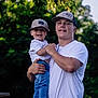 child, toddler, young_man, holding, outdoor, nature, greenery, casual_clothing, smiling, happy, hat, jeans, white_shirt, white_shoes, portrait, family, daylight, trees, fun, people