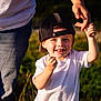 child, toddler, boy, cap, white_tshirt, hand_holding, adult, outdoor, grass, sunlight, smiling, happy, portrait, casual_clothing, nature, summer, person, face, blue_eyes, playing