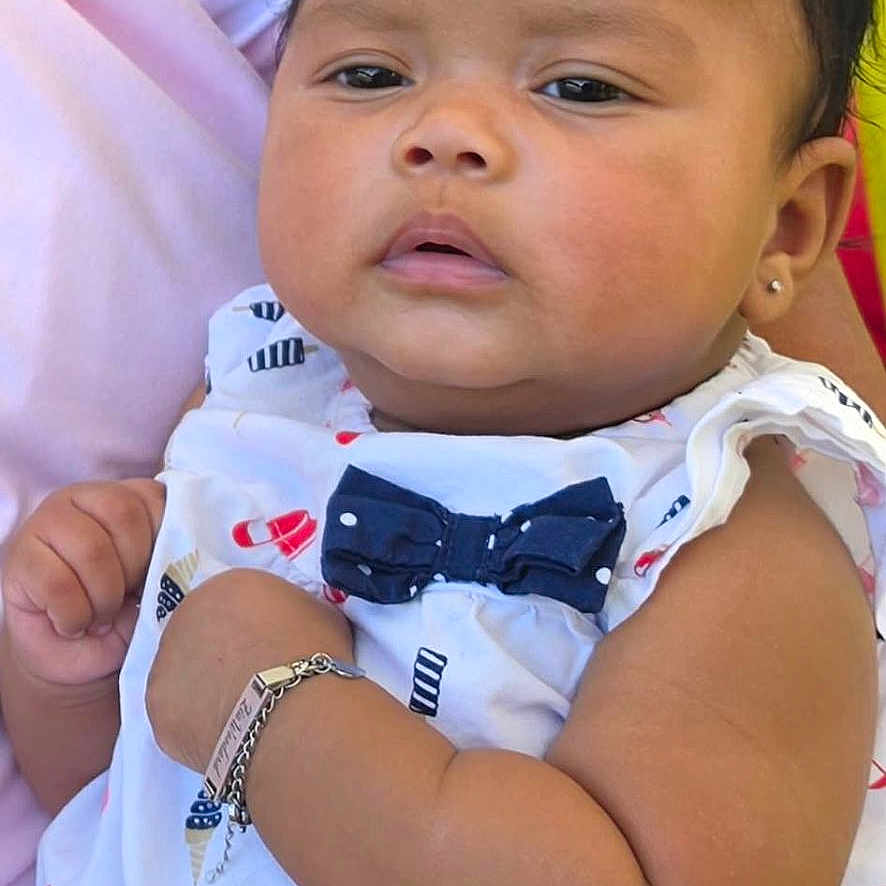 Zia Le'Ashia is registered to the contest to win money with this photo: baby, child, infant, person, outdoor, greenery, clothing, white_dress, bow, bracelet, earring, hand, arm, adult_hand, portrait, cute, serious_expression, holding, skin, head