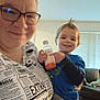 Jeff is registered to the contest to win money with this photo: adult, child, toddler, smile, glasses, bottle, ibuprofen, hairstyle, mohawk, indoor, living_room, couch, window_blinds, clothing, person, holding, happy, family, casual, portrait