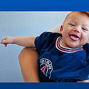Liam-Noha Chalaye a rejoint le concours — aidez-le/la à gagner de superbes lots ! baby, child, smiling, tongue_out, blue_shirt, paris_logo, arm, hand, person, holding, happy, indoor, wall, texture, casual_clothing, portrait, cute, infant, young_child, human