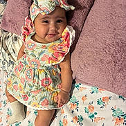 Brianne is registered to the contest to win money with this photo: baby, child, infant, smile, dress, headband, pillow, bedspread, floral_pattern, sock, lying_down, cute, indoors, colorful, soft_texture, person, happy, young, portrait, cozy