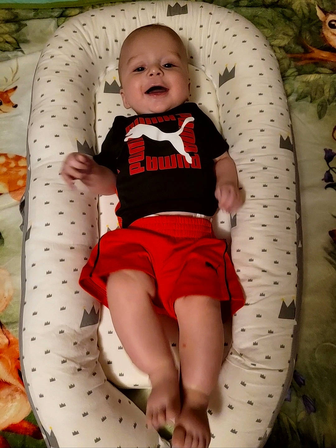 Xander is registered to the contest to win money with this photo: baby, baby_products, baby_toddler_clothing, chair, child, comfort, facial_expression, fun, happy, human_leg, knee, linens, pattern, person, shorts, sitting, sleeve, smile, t_shirt, thigh
