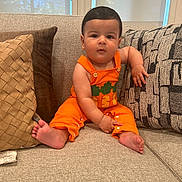 Ryan is registered to the contest to win money with this photo: baby, child, orange_overall, carrot_design, couch, pillows, indoor, cute, infant, sitting, barefoot, home, portrait, young_child, furniture, relaxed, casual, window, natural_light, expression