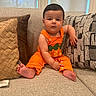 baby, child, orange_overall, carrot_design, couch, pillows, indoor, cute, infant, sitting, barefoot, home, portrait, young_child, furniture, relaxed, casual, window, natural_light, expression