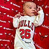 Muhammad is registered to the contest to win money with this photo: baby, infant, onesie, clothing, red_blanket, chicago_bulls, logo, text, sports, basketball, face, expression, hand, lying_down, portrait, cute, indoors, background, closeup, child