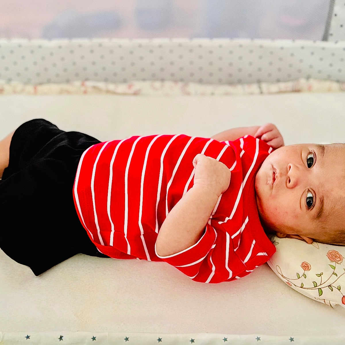 Muhammad is registered to the contest to win money with this photo: baby, infant, child, red_shirt, striped_shirt, black_shorts, lying_down, pillow, floral_pillow, indoor, crib, blanket, face, skin, feet, arms, expression, cute, young, resting