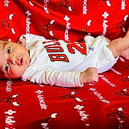 Muhammad is registered to the contest to win money with this photo: baby, infant, newborn, blanket, red, white_clothing, onesie, face, lying_down, cute, small, skin, expression, pattern, text, cartoon, bull, soft, comfort, portrait