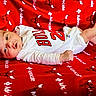 baby, infant, newborn, blanket, red, white_clothing, onesie, face, lying_down, cute, small, skin, expression, pattern, text, cartoon, bull, soft, comfort, portrait