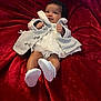 baby, infant, child, white_dress, grey_jacket, white_shoes, red_blanket, soft_texture, smiling, cute, portrait, lying_down, indoors, cozy, warm_clothing, small_hands, happy, baby_face, plush, comfortable