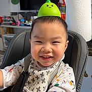 Kash is registered to the contest to win money with this photo: child, toddler, smile, high_chair, bib, toy, green, face, indoors, kitchen, happy, clothing, short_hair, seat, playful, colorful, background, expression, person, cute