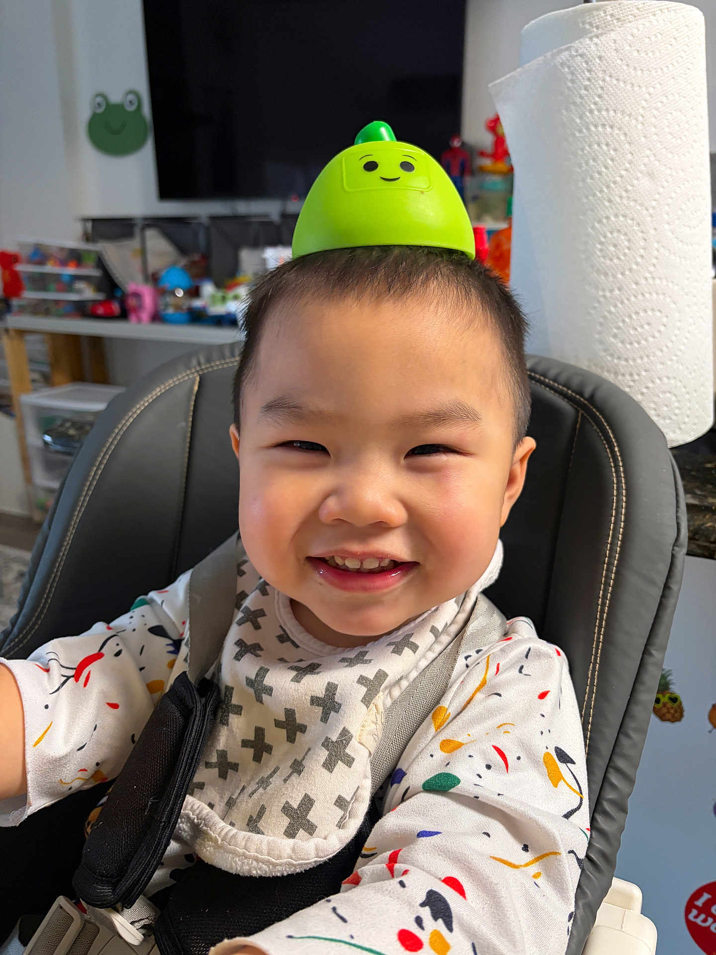 Kash is registered to the contest to win money with this photo: child, toddler, smile, high_chair, bib, toy, green, face, indoors, kitchen, happy, clothing, short_hair, seat, playful, colorful, background, expression, person, cute