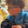 child, boy, glasses, blue_jacket, black_jacket, fleece, hair, side_profile, hands, table, indoor, window, elderly_person, blurred_background, outdoor_scene, seated, casual_clothing, portrait, daylight, pensive