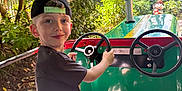 Gabriel participe au concours pour gagner de l'argent avec cette photo : child, boy, cap, black_tshirt, green_vehicle, steering_wheel, amusement_ride, outdoor, trees, sunlight, seat, playful, nature, park, smile, shorts, daylight, leafy, fun, recreation