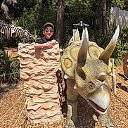 Gabriel a rejoint le concours — aidez-le/la à gagner de superbes lots ! child, boy, sunglasses, cap, dinosaur, triceratops, statue, rock, outdoor, sunlight, trees, woodchips, skeleton, t_rex, nature, fun, smile, park, adventure, play