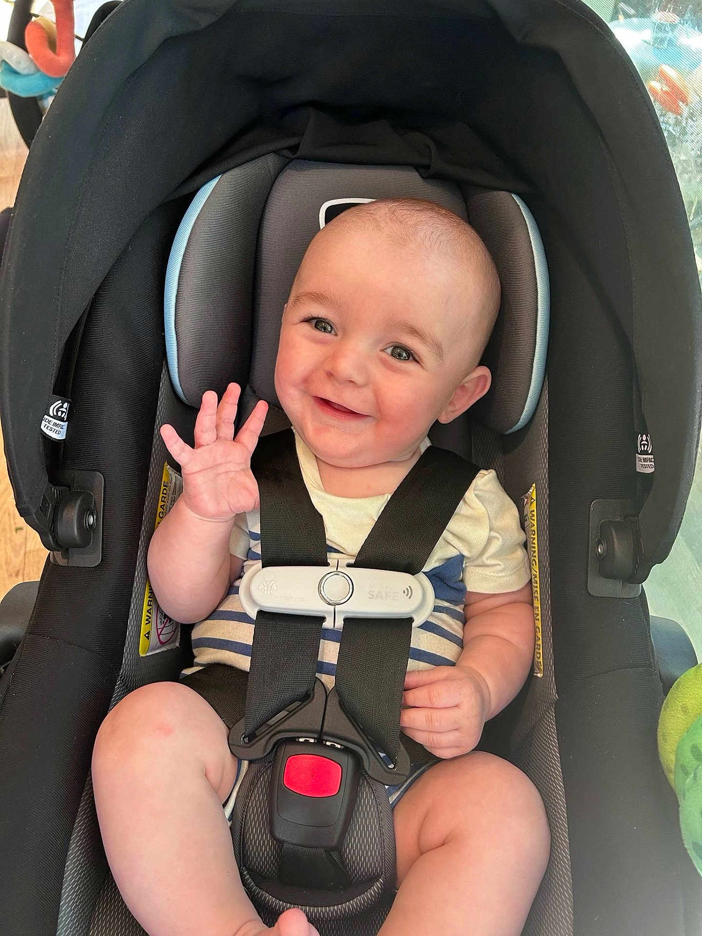 Wesley is registered to the contest to win money with this photo: baby, car_seat, smiling, waving, infant, child, seatbelt, safety_harness, indoor, cute, happy, person, head, face, clothing, striped, skin, arm, leg, toy