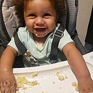 Reign is registered to the contest to win money with this photo: baby, child, high_chair, smiling, eating, food, crumbs, messy, mealtime, portrait, face, curly_hair, infant, sitting, indoors, harness, tray, happy, playful, family