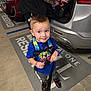 adult, backpack, blue_shirt, boy, car, casual_clothing, child, concrete, footwear, gray_pants, hand, parking_lot, playful, reserved_parking, smile, standing, suv, tattoo, urban, vehicle